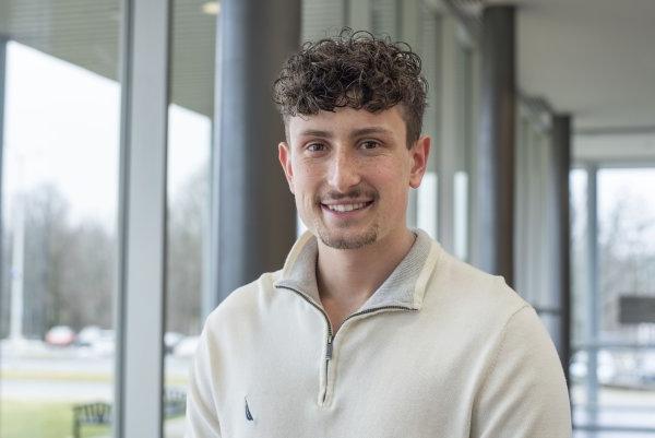 Conner Crossley, in white zip-up pullover, stands by a window in Bull Hall and smiles.